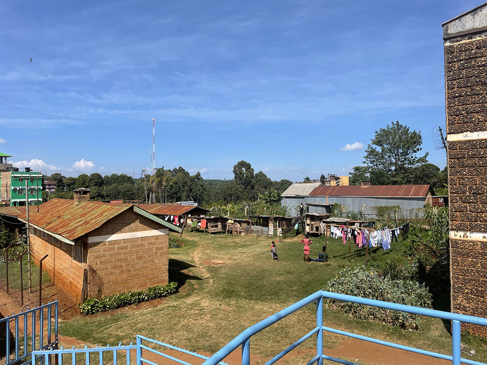 The image shows a rural area with a collection of small houses, some with visible laundry hanging outside. There is an open field adjacent to the houses and a clear sky overhead.