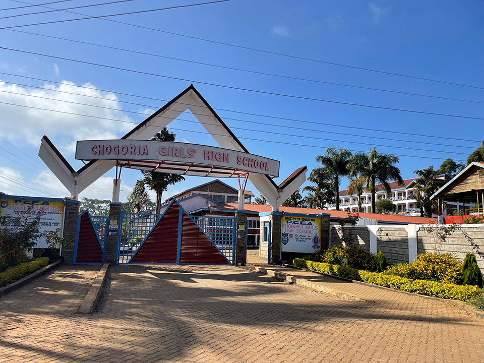 The image depicts a school entrance with a sign that reads 'Chinoka High School,' located in a sunny, tropical environment.