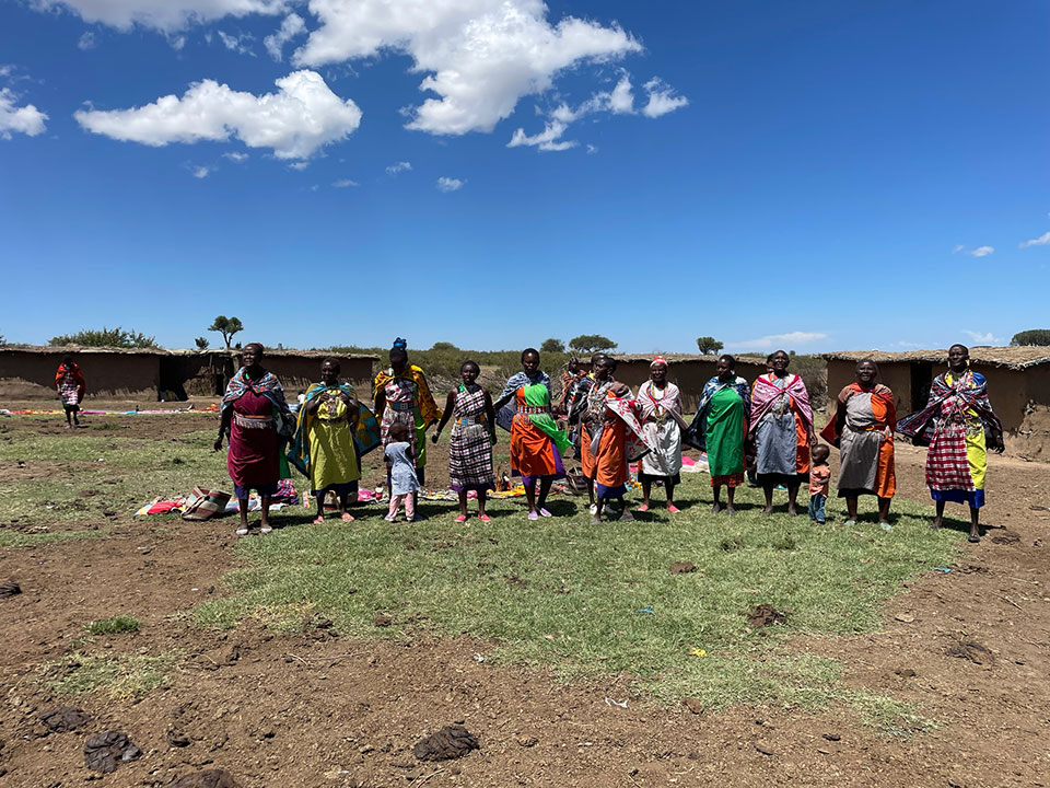 The image shows a group of people standing on an open field, under a clear blue sky with some clouds. They appear to be in the middle of a gathering or event, possibly related to cultural or traditional activities, as suggested by their attire and the presence of colorful blankets on the ground.