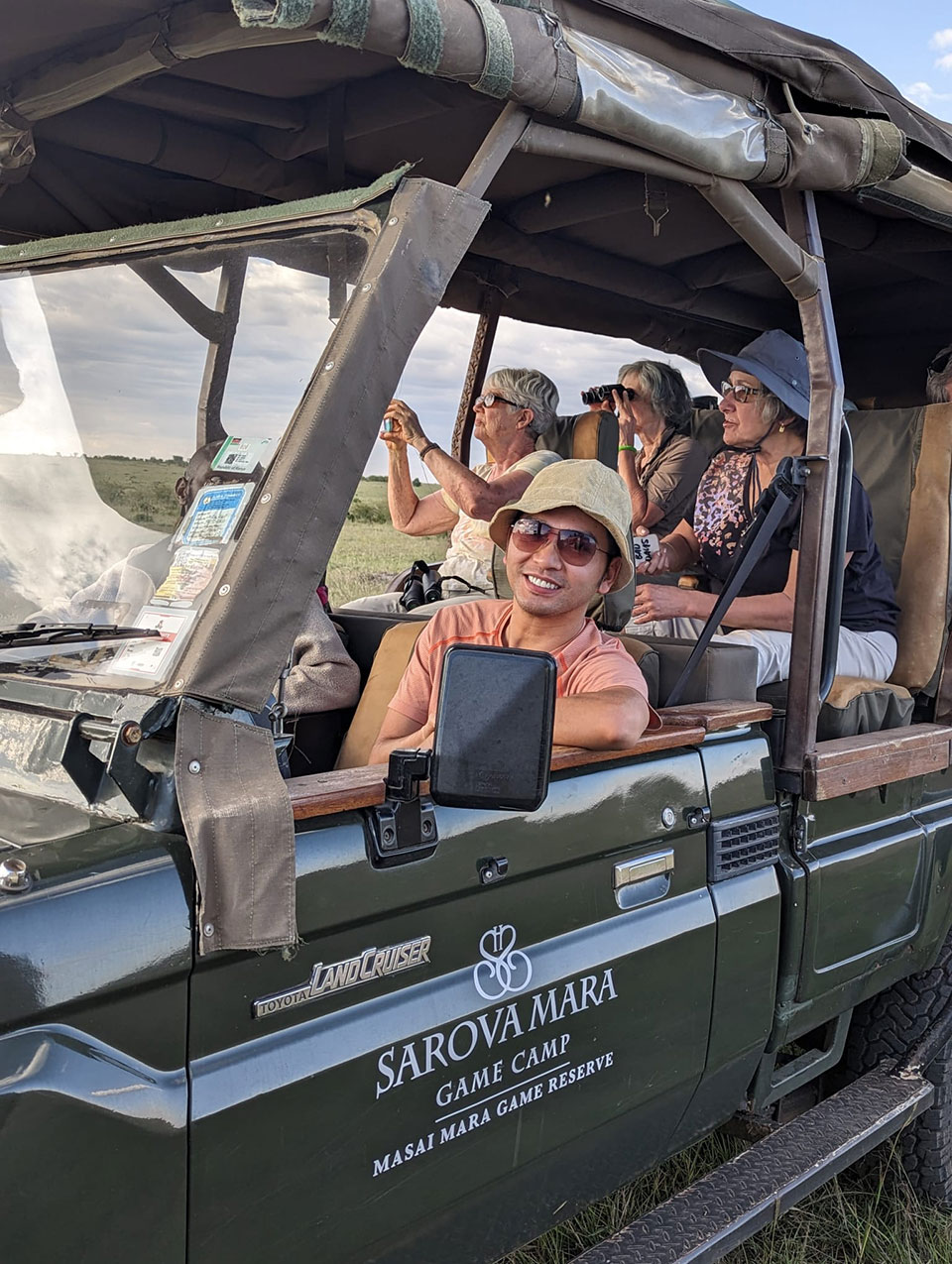 This image depicts a group of people enjoying a safari tour in an open-top jeep, with one individual at the wheel and others seated around. The vehicle is parked on a dirt road, and the landscape suggests a natural environment, possibly during sunset or early evening, given the soft lighting.