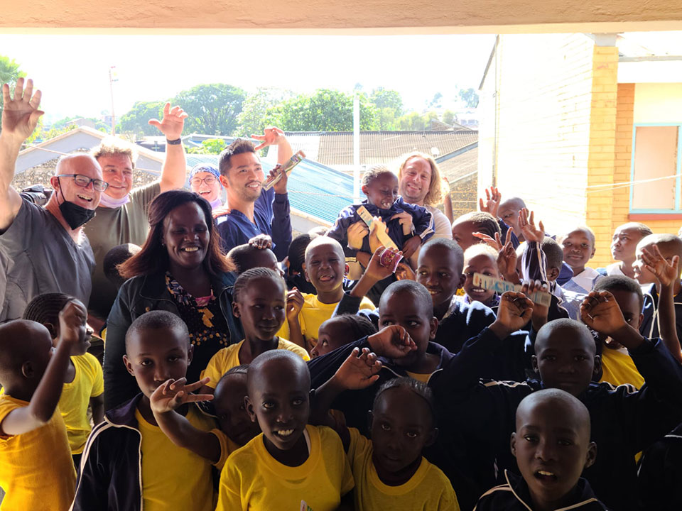 A group of people, including children, are gathered in a room with a large window. They appear to be posing for a photo with their arms raised and hands open, suggesting they are celebrating or welcoming someone. The setting looks like a community center or school, indicated by the presence of uniforms on some of the individuals.