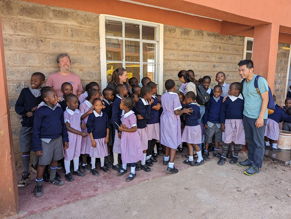 The image shows a group of people, including children and adults, posing for a photograph in front of a building. A man stands to the side, overseeing the gathering.