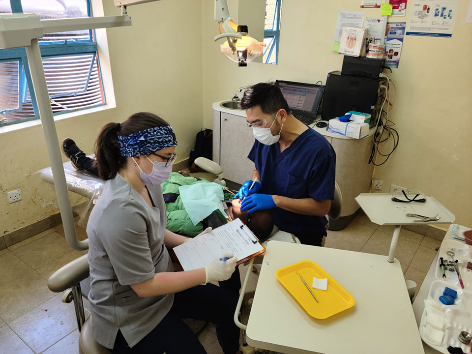 A medical professional in a mask and protective eyewear attends to a patient with paperwork, in an office setting.