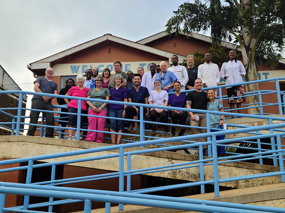 The image shows a group of people standing on a concrete platform in front of a building with the sign 'Welcome to the Village.' They appear to be posing for a photograph.