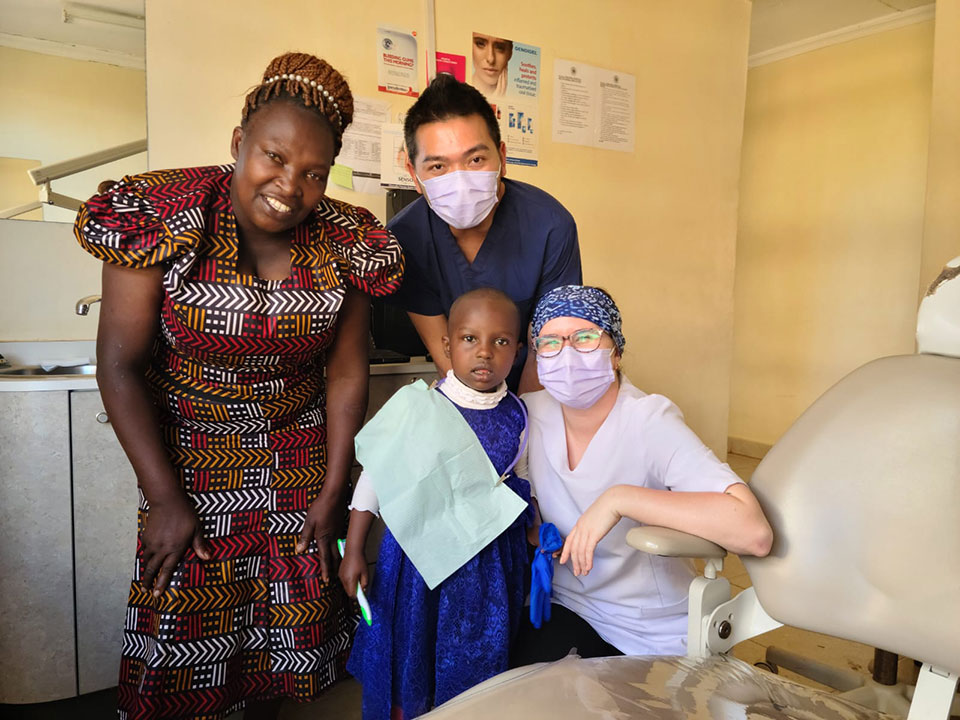 A group of people, including a man in a white shirt and tie, gathered around a child wearing a surgical mask, posing for a photo in what appears to be a dental or medical setting.