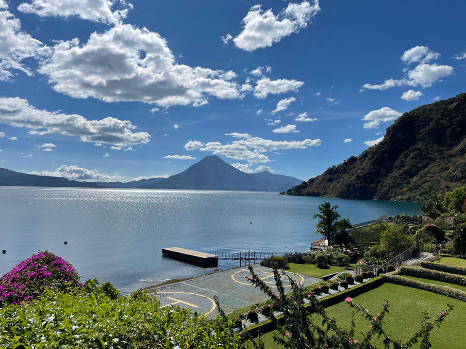 The image depicts a serene lakeside view with a clear blue sky, a large body of water reflecting the sunlight, and a majestic mountain in the distance.