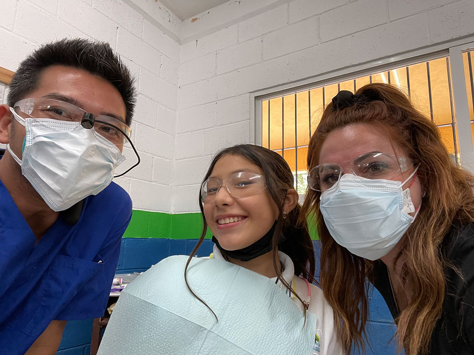 The image shows a group of people in what appears to be a medical or dental setting, with one individual wearing a face mask and smiling at the camera.