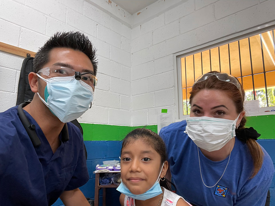 The image shows three individuals in a room, with one person wearing a surgical mask and another two posing for the photograph. They are standing in front of a wall, which has some text on it, but the text is not clearly legible.