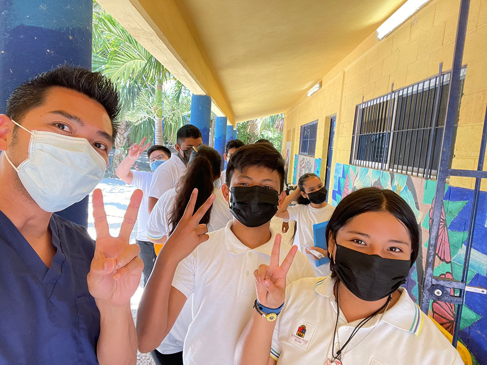 In the image, there is a group of people posing for a photo in front of a school building. They are wearing masks and making peace signs with their hands. The man on the left appears to be a medical professional, standing slightly apart from the group.