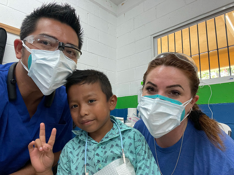 A group of healthcare professionals, including a man and two women, posing in front of a medical facility with a child wearing a surgical mask.