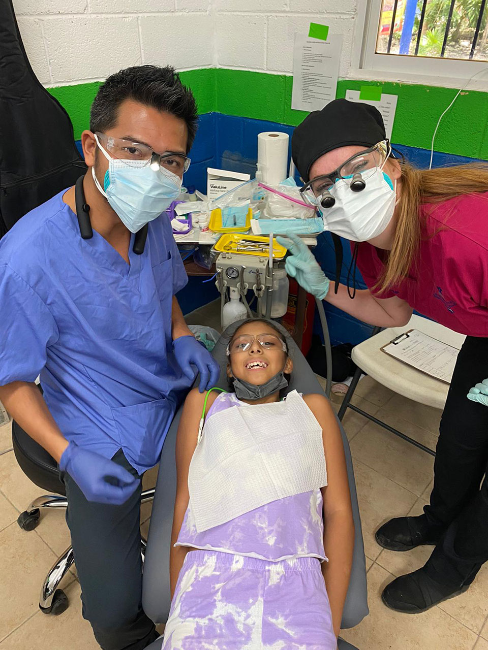 In the image, two individuals are in a medical setting, with one person seated on a chair and another standing nearby. The person seated appears to be receiving dental care or treatment, as indicated by the presence of dental instruments and tools around them. They are wearing a purple garment that covers their body from the chest down, suggesting some form of medical procedure or recovery process. The individual standing is dressed in typical dental office attire, including a white coat, blue scrubs, and a face mask, indicating they may be a dental professional providing care. There are other individuals present as well, one of whom is wearing a black apron and a face mask, possibly another medical professional or staff member. The environment includes a chair with wheels, a desk with various items, and a large window that lets in natural light.