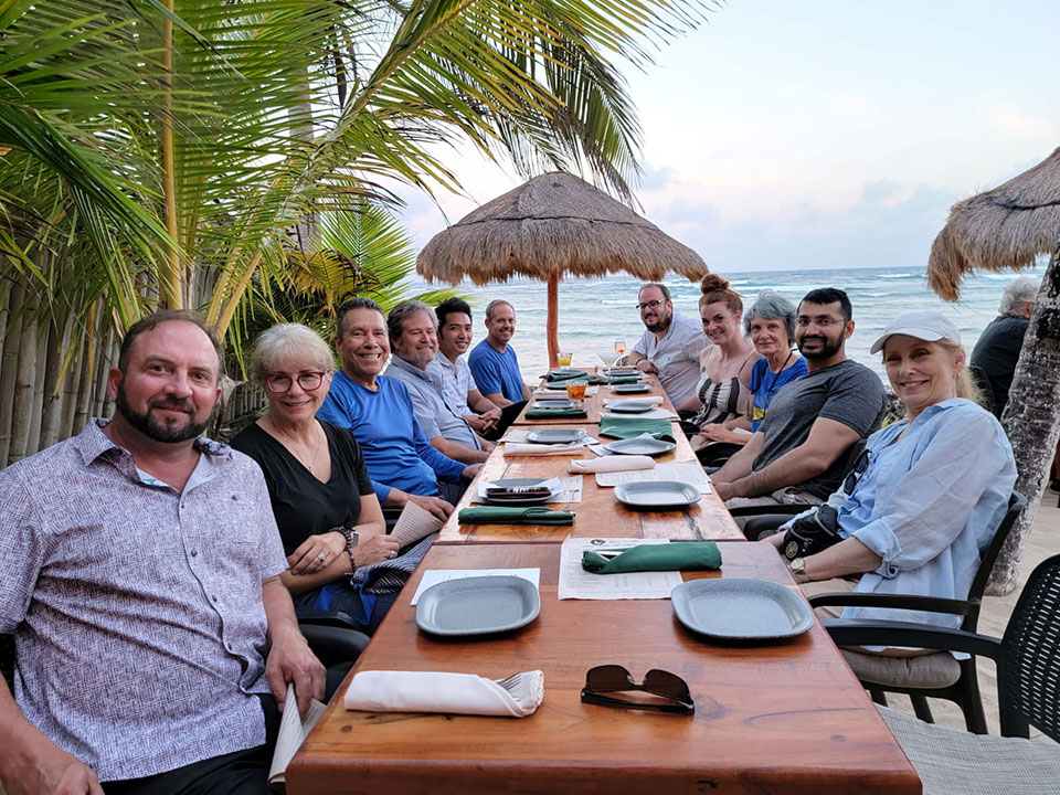 A group of people sitting at a dining table with plates and cutlery, posing for a photo in front of a beachfront restaurant with thatched roofs.
