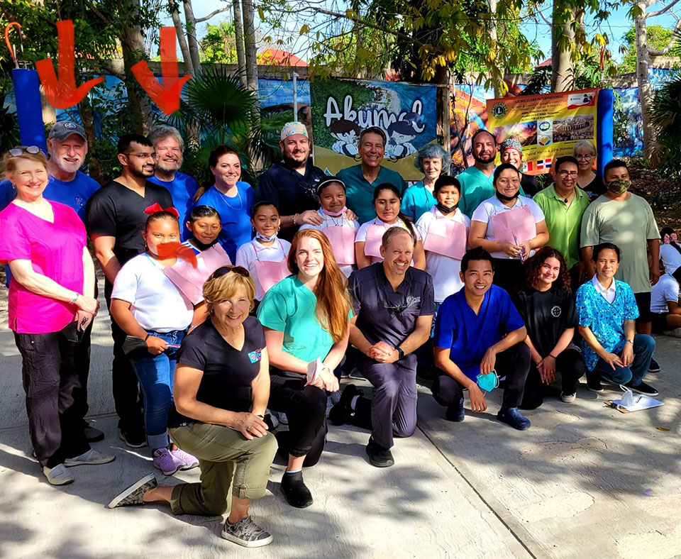 The image is a photograph of a group of people posing for a photo outdoors. They are standing on a paved surface, and some individuals are wearing name tags with visible text.