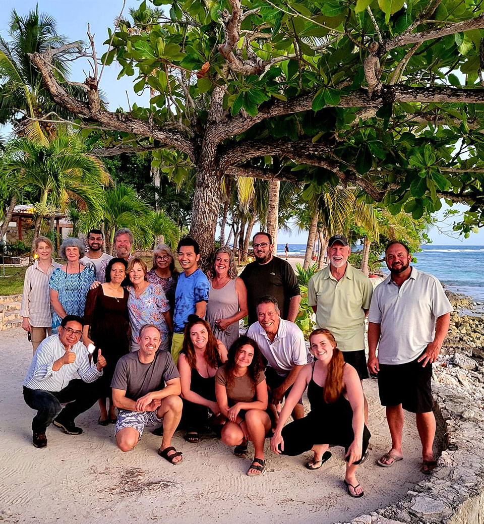 A group of people posing for a photo on a beach, with palm trees and a clear sky in the background.