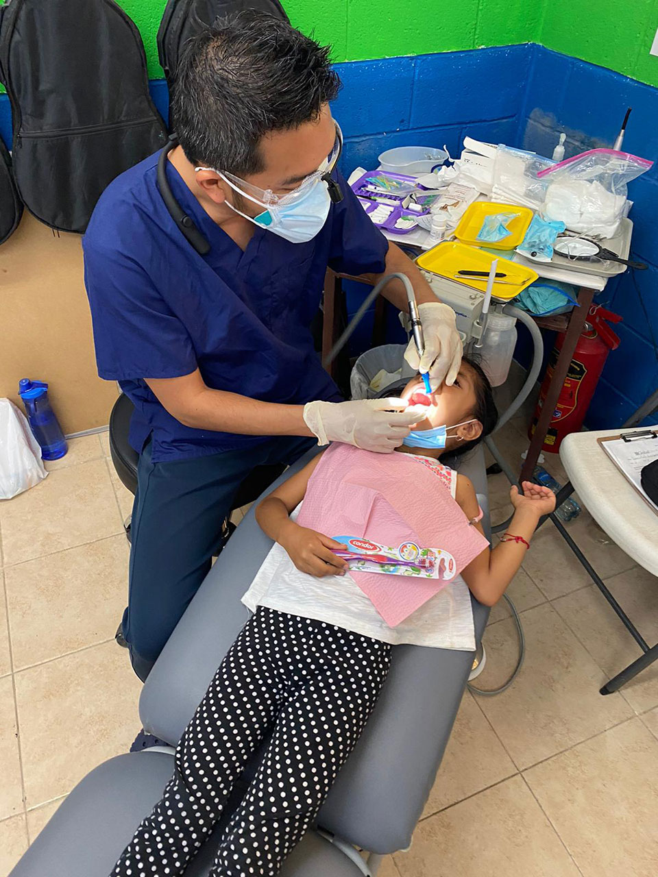 A dental hygienist in a blue uniform is performing a dental cleaning on a young child in a chair, with the child wearing a pink bib and lying down.