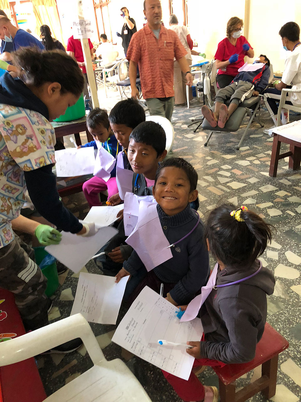 This is a photograph of a group of children sitting in front of an adult, all wearing face masks and engaging with papers or drawings on their laps. They are indoors, likely in a classroom setting, as indicated by the presence of chairs, desks, and what appears to be a medical or dental professional assisting them.