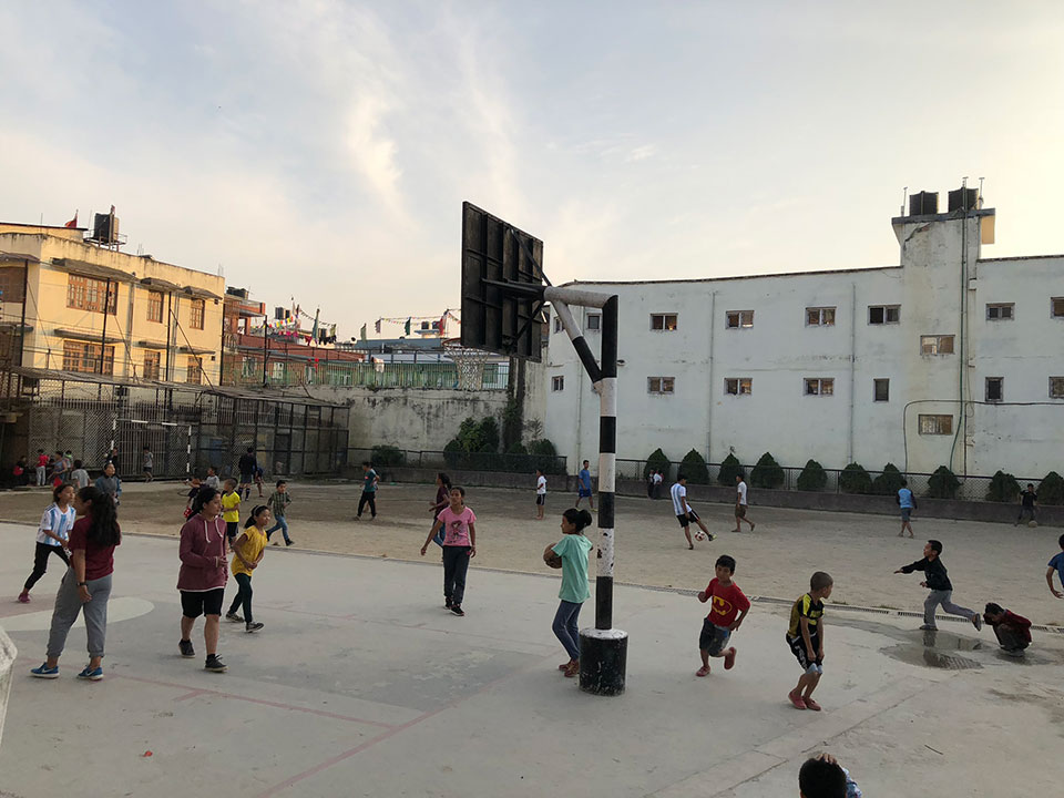 The image shows a basketball court with people playing, an outdoor setting at dusk with a clear sky, and a building in the background.