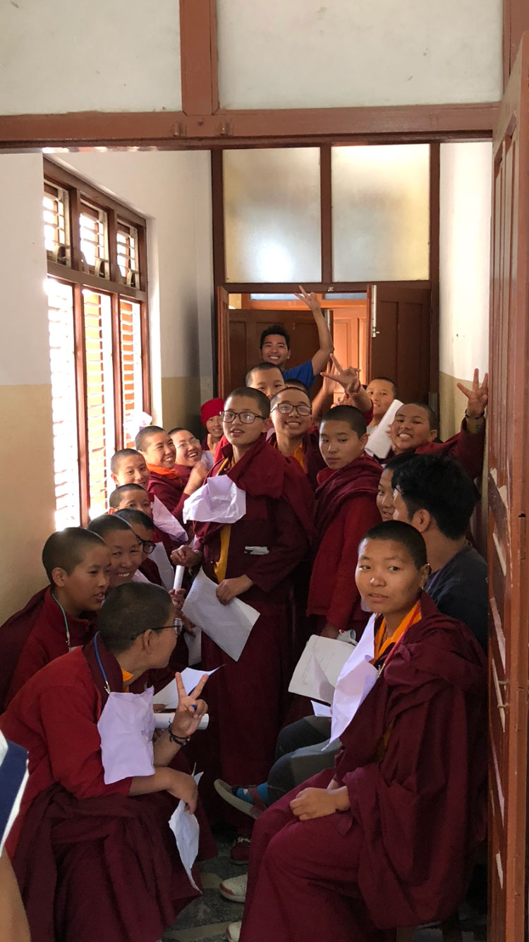 The image depicts a group of people, possibly monks or students, gathered in an indoor setting with papers and a booklet visible. They are seated on the floor, facing towards the camera, with some individuals holding up their papers.