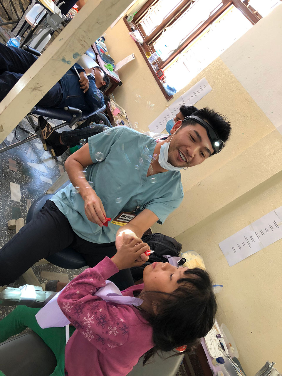 The image shows a man in a medical uniform, likely a dentist or oral surgeon, interacting with a child. He is holding what appears to be a small object, possibly a toothbrush or dental tool, and the child is receiving it. They are both indoors, and there are other people in the background who seem to be engaged in various activities. The setting suggests an educational or community event focused on oral health care.