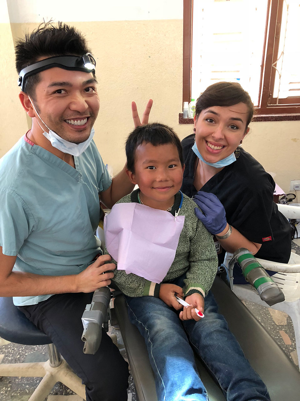 In the image, there is a group of three people in a dental setting. A dentist is sitting on a chair with a child and another person standing behind him. They are all wearing face masks and appear to be engaged in dental care activities. The environment suggests a professional dental clinic.