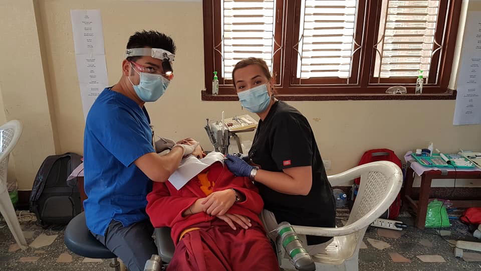 Medical professionals in a makeshift setting, performing dental work on a patient, with equipment and personal protective gear visible.