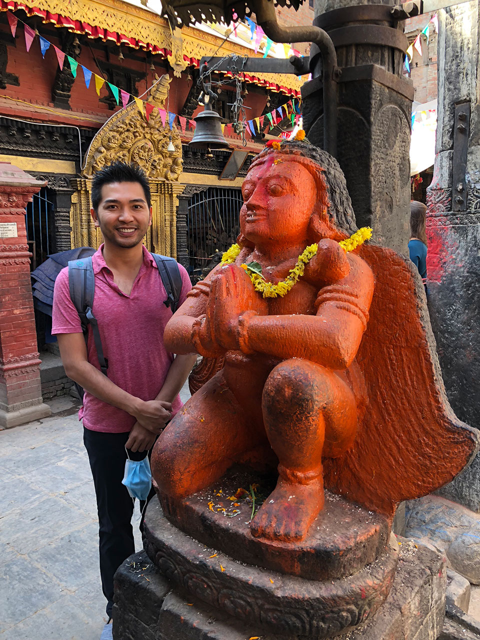 The image shows a person standing next to a large, red, ornate statue of a seated figure that appears to be a deity, possibly in an Indian cultural context, with the backdrop featuring traditional architecture and colorful flags.