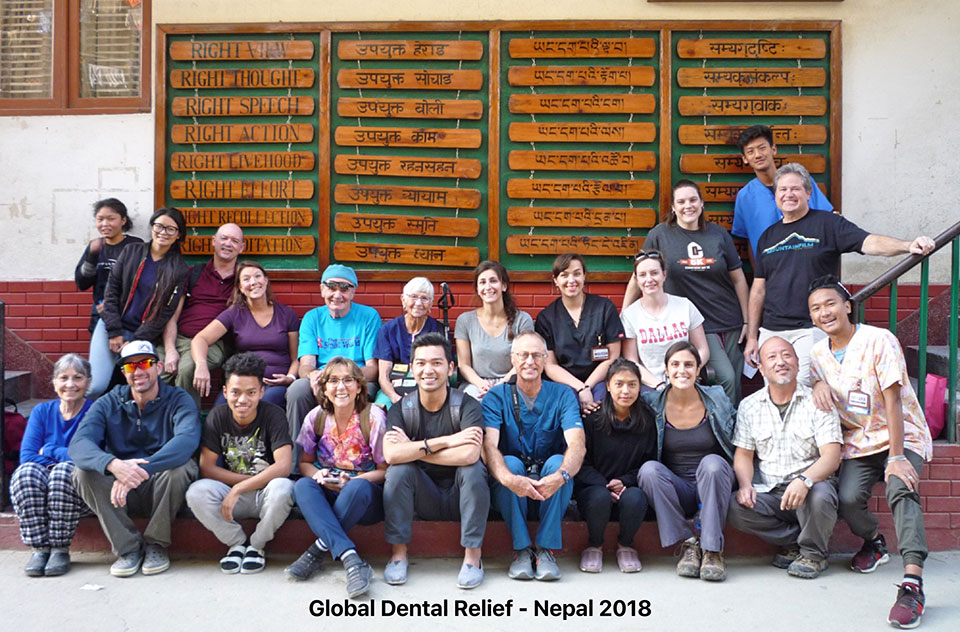The image shows a group of people posing in front of a wall with boards displaying names and information, likely at a dental clinic or educational institution. They are standing on steps, smiling and looking towards the camera.
