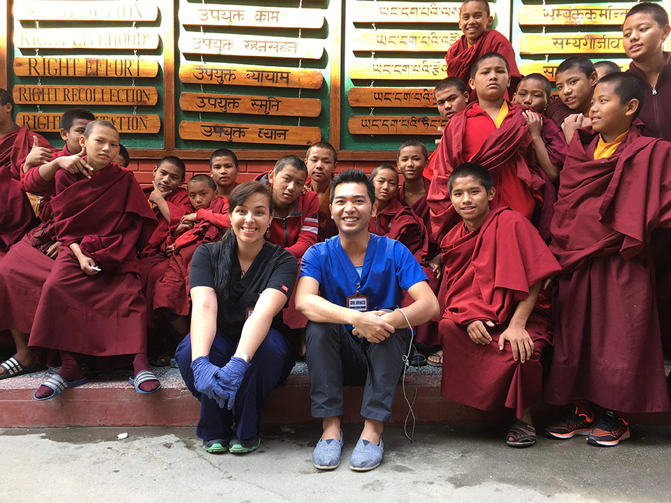 A group of people, including a woman in blue and a man in white, posing with a sign featuring writing in multiple languages, standing together outdoors.