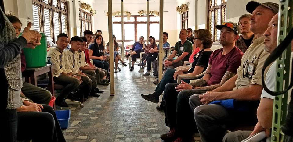 The image shows a group of people, including individuals who appear to be in uniforms, seated on benches inside what seems to be a public building with a high ceiling.