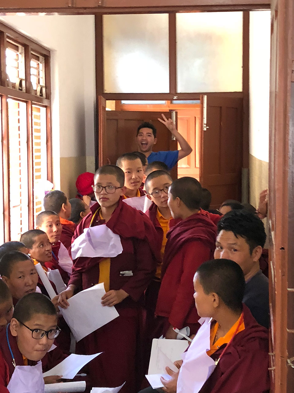 An image of a group of people, likely monks or a similar group, gathered in a room with a man standing at the doorway. They are seated and appear to be engaged in an activity involving papers or documents.
