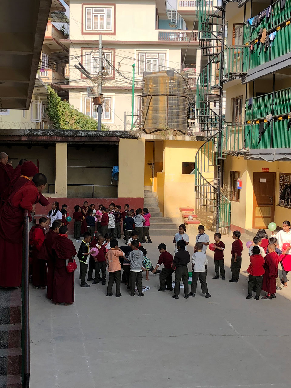 The image shows a group of people, including adults and children, gathered in an outdoor area with buildings in the background. They are standing on a concrete surface, and some individuals appear to be interacting or engaging with each other. The setting suggests it could be a school or community event.
