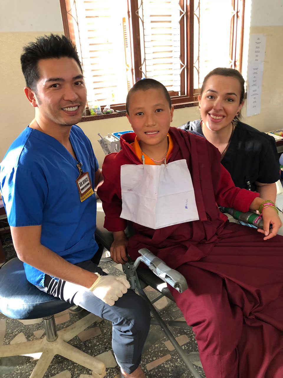 The image shows three individuals in a room, where one person is sitting on a chair with a medical mask covering their mouth and nose, another person is standing behind them, and the third person is partially visible at the front of the frame. They are all inside a room that appears to be a healthcare setting, as indicated by the presence of medical equipment and the professional attire of one individual.