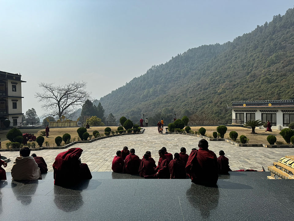 The image shows a group of people seated in front of a large, open space with a clear sky and mountains in the background. They are dressed in traditional monastic robes, suggesting they may be Buddhist monks or nuns, gathered for a ceremony or event at a religious site.