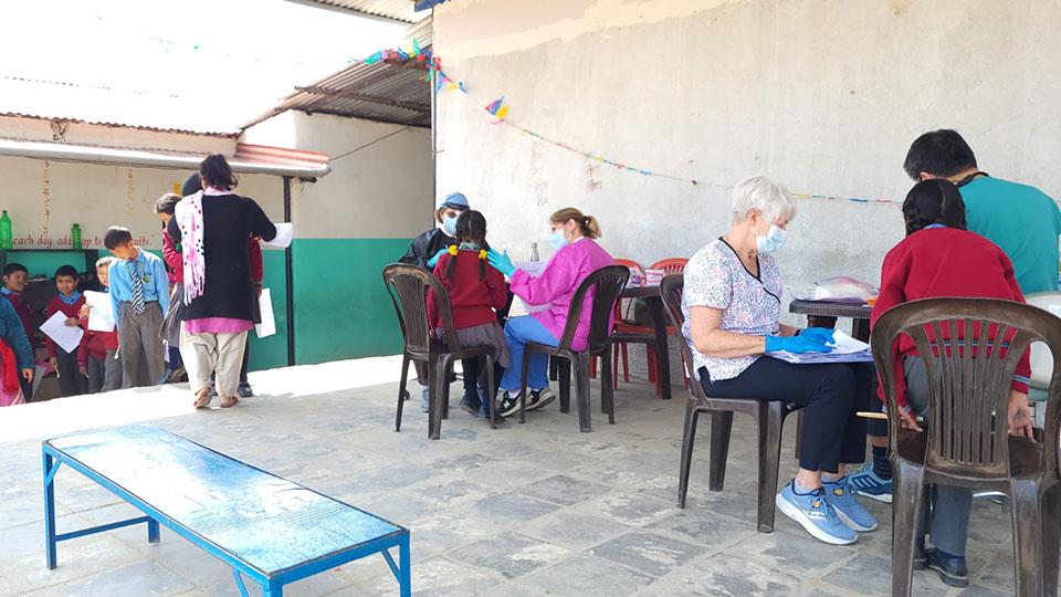 The image shows an indoor setting with a group of people, some seated at tables and others standing. There are chairs, tables, and individuals engaged in various activities such as writing or reading. A person is wearing a face mask, and the environment appears to be a communal space, possibly for learning or socializing.