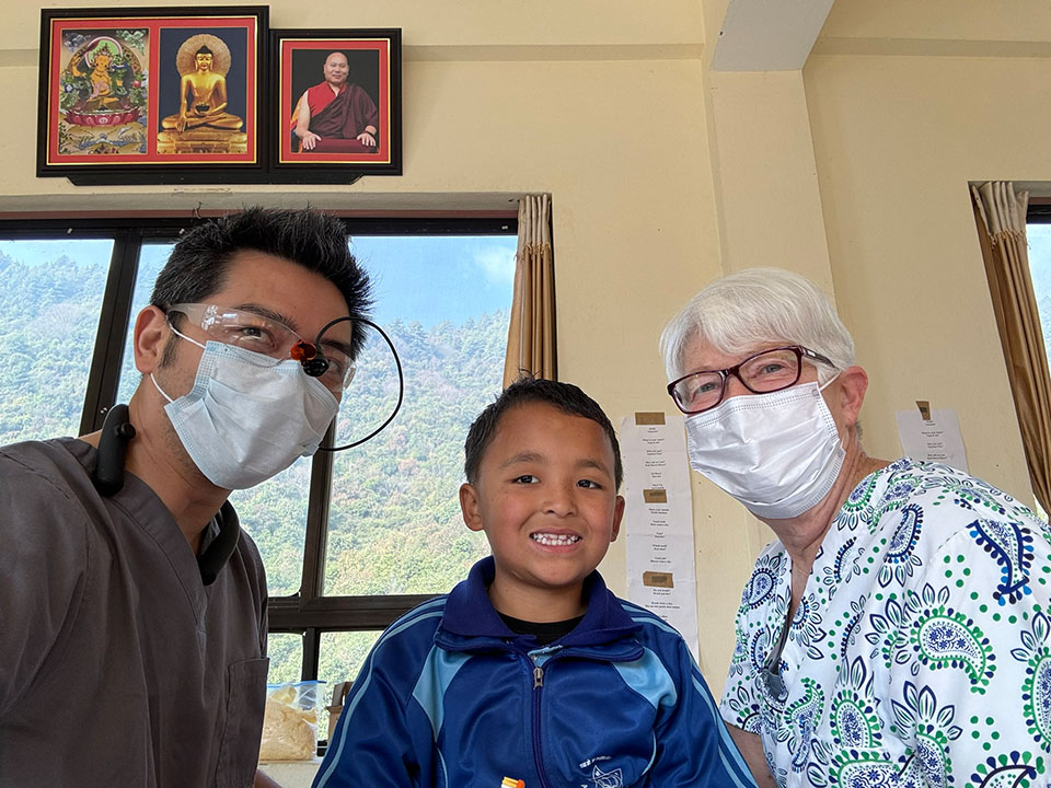 The image shows three individuals, two adults and a child, wearing face masks and posing in front of a window with a view of mountains. They are indoors, possibly in a room with a religious or cultural theme, as indicated by the presence of a framed picture on the wall behind them.