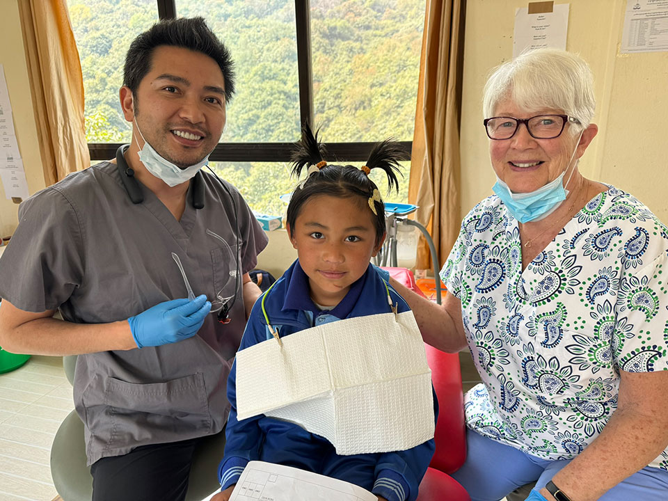 A photograph of three individuals, one standing and two seated, in a room that appears to be a dental or medical office, with the person in the foreground holding an object that looks like a dental implant.