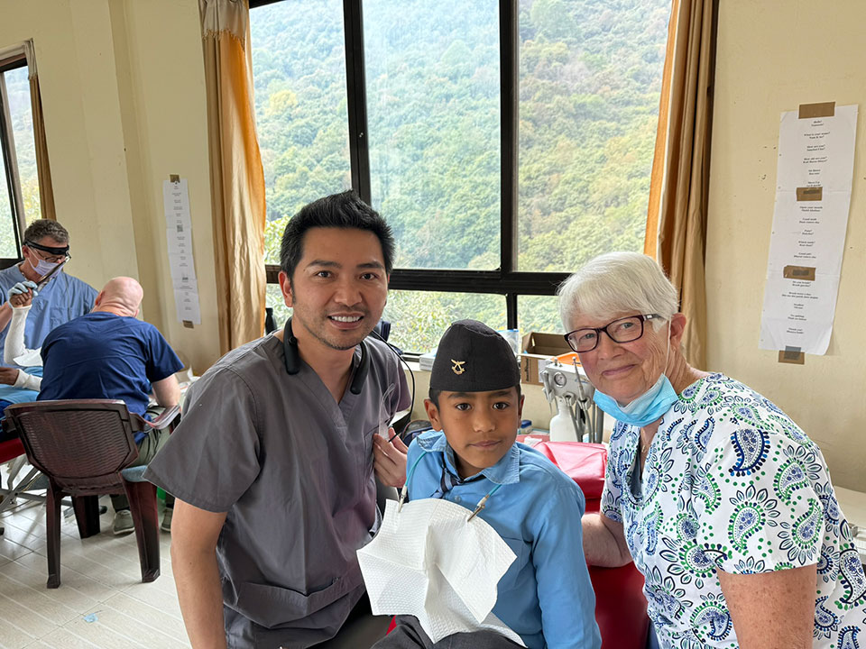 The image shows a group of people in a room with medical equipment, including what appears to be a patient lying on a bed. A man is standing next to the bed, smiling at the camera, while a woman stands beside him, also smiling and posing for the photo. They are both dressed in scrubs, suggesting they may be healthcare professionals. The setting seems to be a medical facility or a room designated for medical care.