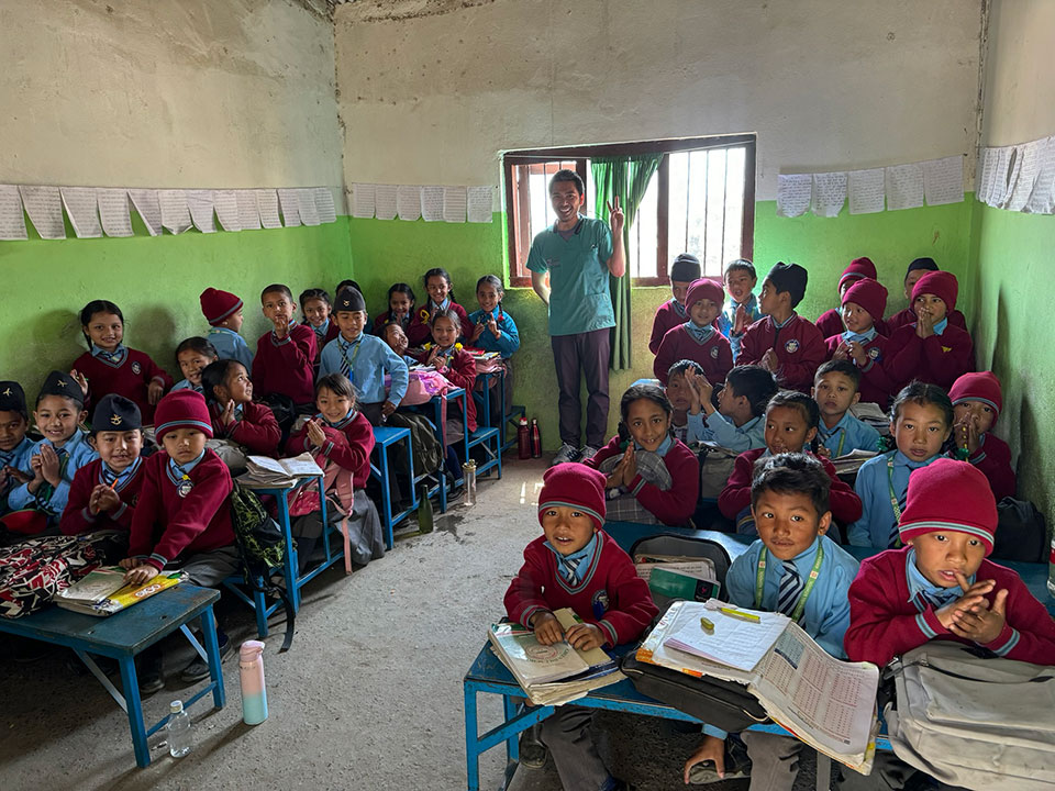 The image depicts a group of students in a classroom setting, with a teacher standing at the front of the room.