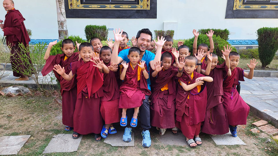 The image shows a group of children in traditional Buddhist attire posing for a photo with an adult, likely a monk, behind them. They are outdoors, and the setting appears to be a monastery or temple courtyard.