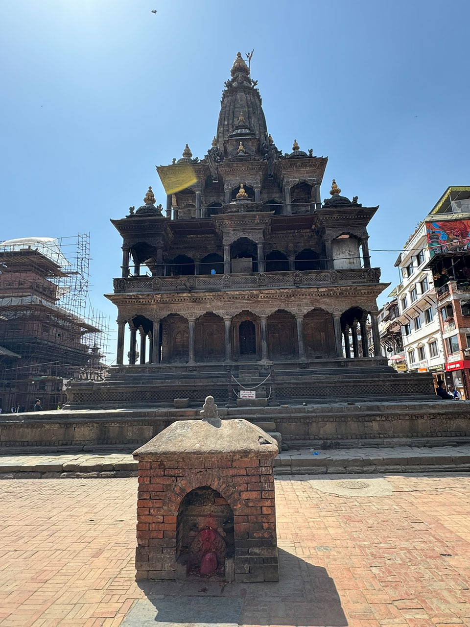 The image depicts an ornate, multi-tiered structure that resembles a traditional Indian temple with intricate architectural details, situated in a courtyard within what appears to be a historic urban setting.
