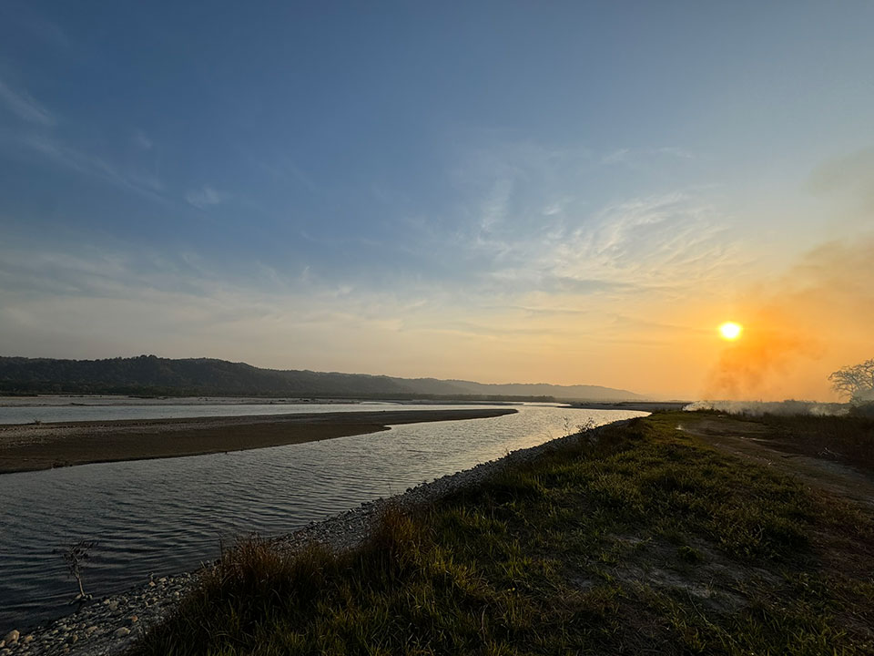 The image depicts a serene landscape at sunset, featuring a calm river with a bank lined by rocks and vegetation, leading to a distant horizon where the sky meets the water.
