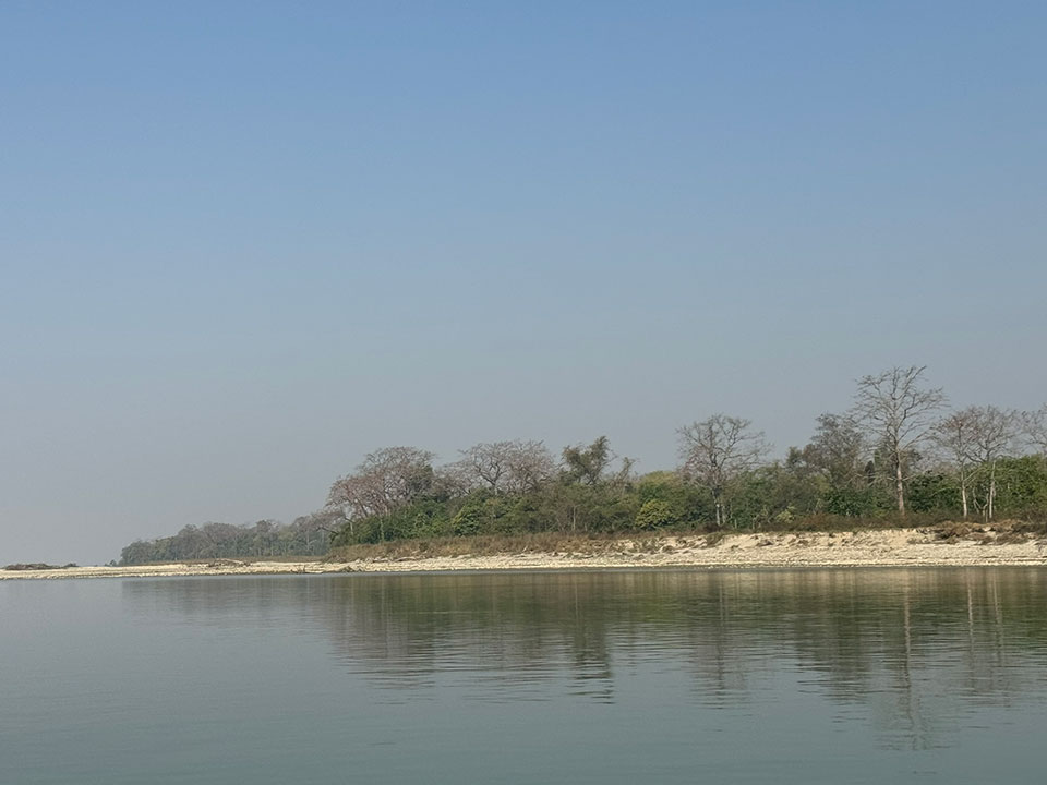 The image shows a serene riverbank with a clear blue sky, calm water reflecting the hazy horizon, and a sandy bank lined with vegetation.