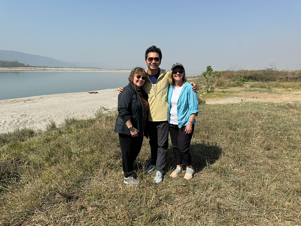 The image shows two individuals posing outdoors, with a body of water in the background and a clear sky above.