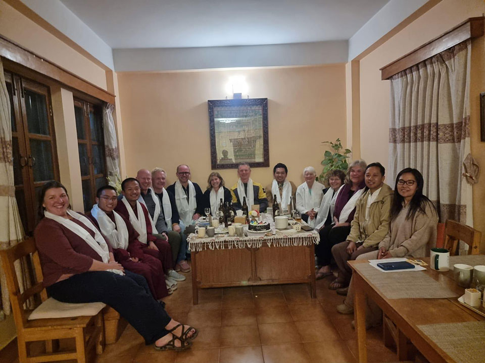 A group of people gathered around a dining table in what appears to be a living room, with some members wearing white scarves and others seated on chairs, all seemingly engaged in conversation or an event.