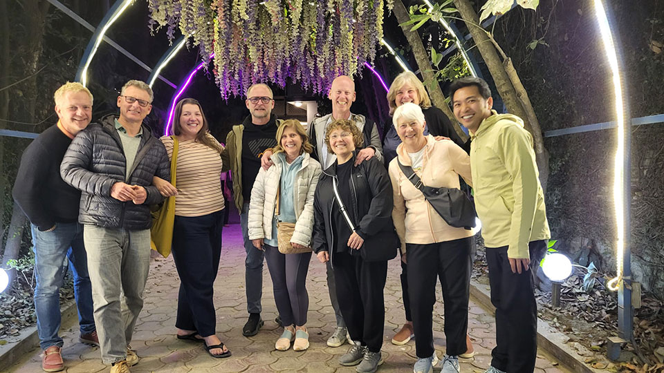The image shows a group of people posing for a photo in front of a purple flower archway at night.