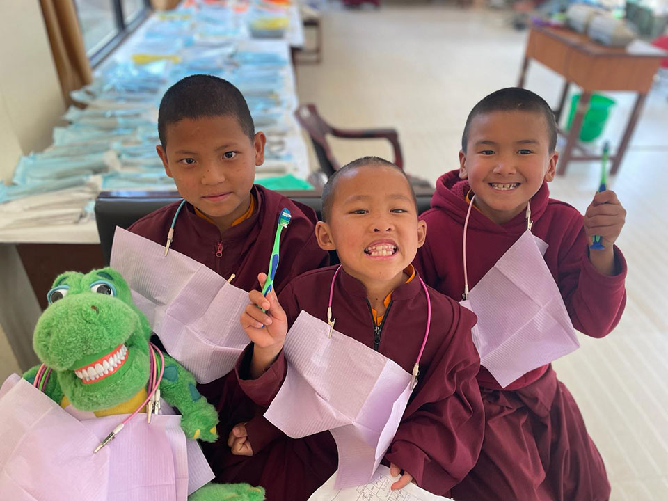Three young boys in traditional monastic robes, each holding a toothbrush and smiling at the camera, with one boy also holding a stuffed toy.