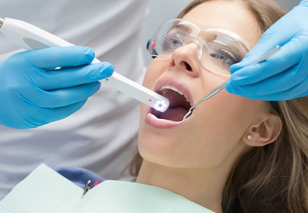 The image shows a woman in a dental chair receiving dental treatment. She is wearing a blue surgical gown, and there are medical devices around her neck. A dental professional is using a laser device to perform the procedure.