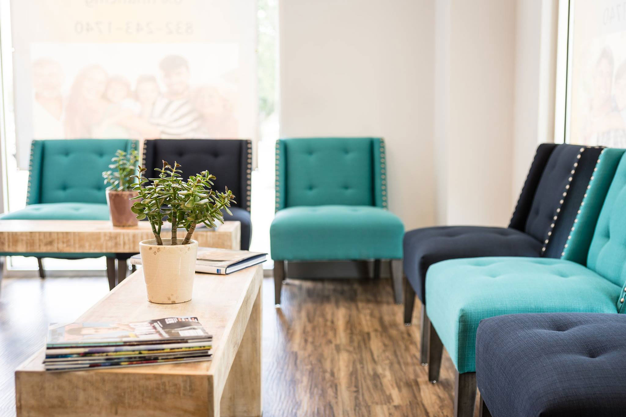 The image shows a modern waiting room with blue and black upholstered furniture, including chairs and sofas, arranged around a central coffee table. A potted plant is visible on the table. The flooring appears to be a light-colored wood or laminate, and the walls are painted in neutral tones. There is no text present in the image.