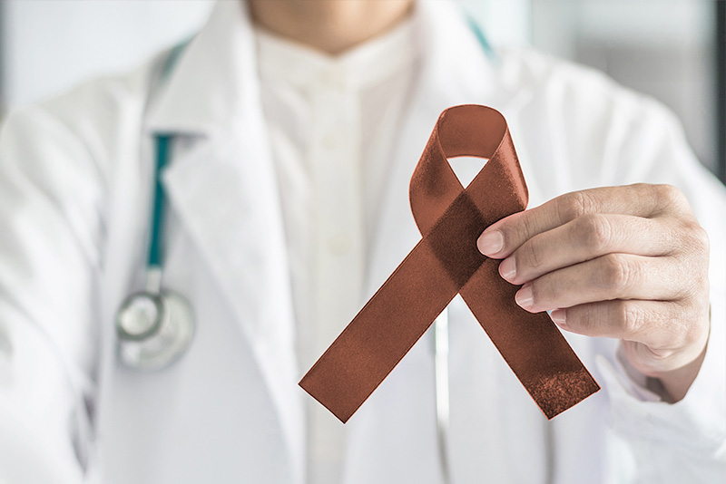 The image is a photograph of a healthcare professional, likely a doctor or nurse, holding up a red ribbon with a symbol for cancer awareness.