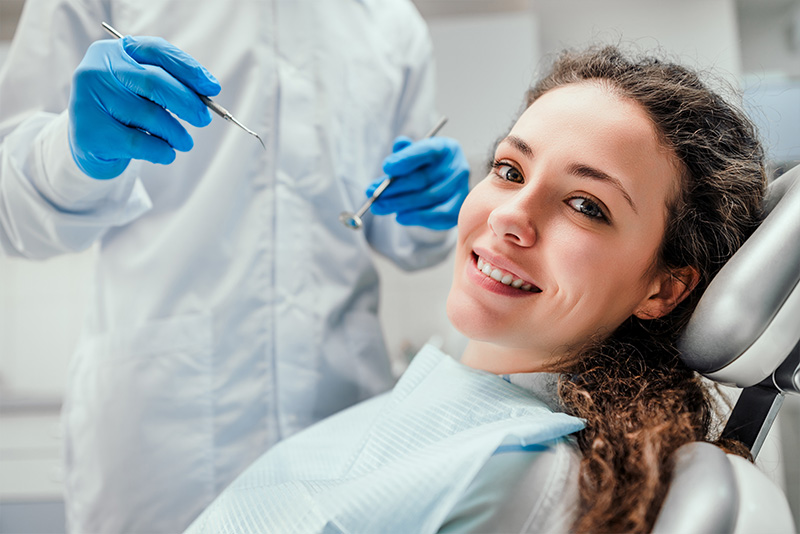 A woman in a dental chair is smiling at the camera, with a dental hygienist performing a procedure on her teeth.